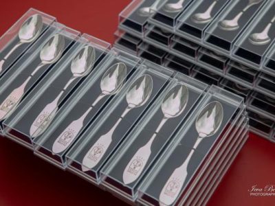 Iesaiņotas sudraba karotes, sakrautas rindās un kaudzēs uz sarkana fona. Katra karote atsevišķā caurspīdīgā plastmasas kastītē ar melnu ieliktni. Uz to rokturiem iegravēts brieža galvas simbols un teksts "SVEICAM VECĀKUS".