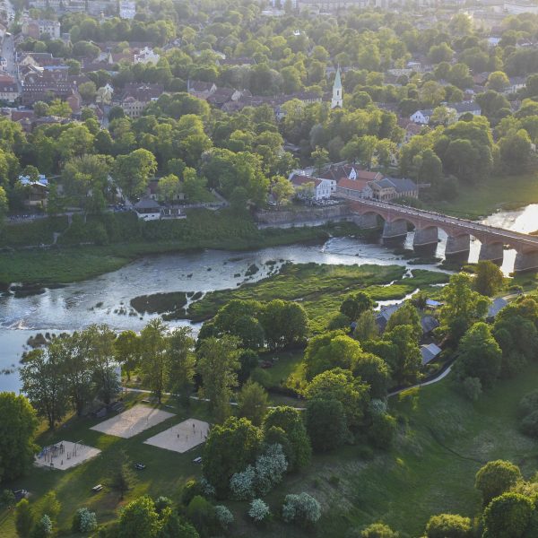 Skats no augšas uz Kuldīgas veco ķieģeļu tiltu, kas šķērso Ventas upi. Upes krastos izvietojusies pilsēta ar ēkām un baznīcas torni, ko ieskauj bagātīgi zaļi koki. Uz upes virsmas labajā pusē atspīd saule, un kreisajā krastā redzami smilšu laukumi un rotaļu laukums.