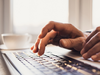 Rokas drukā uz klēpjdatora tastatūras, fonā kafijas tase pie gaiša loga.