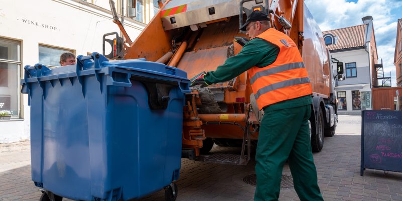 Oranžā atstarojošā vestē tērpies SIA Kuldīgas komunālie sētnieks stumj lielu zilu atkritumu konteineru pie oranžas atkritumu savākšanas mašīnas. Fonā redzamas vecpilsētas ēkas.