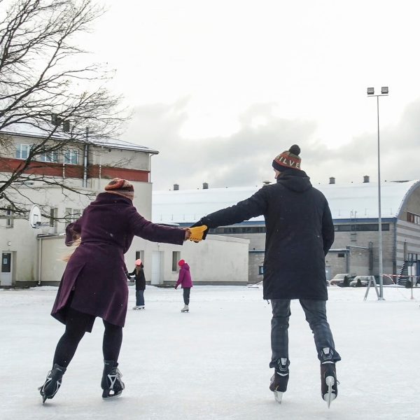 Pāris, turoties pie rokām, slido āra slidotavā ziemas dienā. Vīrietis valkā cepuri ar uzrakstu "LV", bet sieviete cepuri ar uzrakstu "SILVE". Fonā redzamas ēkas un citi slidotāji. Kuldīgas slidotava.