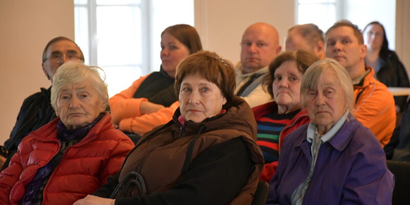 Cilvēku grupa, galvenokārt gados vecāki cilvēki, sēž iekštelpās, klausoties vai vērojot priekšā notiekošo. Priekšplānā redzamas trīs gados vecākas sievietes: viena ar baltiem matiem sarkanā jakā, otra ar brūniem matiem brūnā vestē, trešā ar sirmiem matiem violetā jakā.