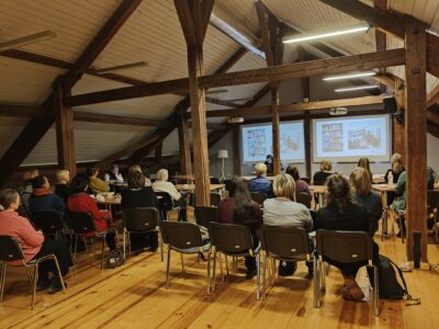 Bēniņu telpa ar atsegtām koka sijām, kurā pieaugušo grupa sēž krēslos, vērojot prezentāciju. Priekšā redzami divi projektoru ekrāni, kuros attēloti grāmatu plaukti un cilvēki, kas lasa vai strādā pie galdiem.