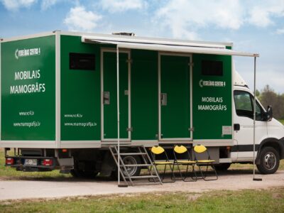 Zaļi-balts kravas auto ar numura zīmi HK-349, uz kura sāniem rakstīts "VESELĪBAS CENTRS 4" un "MOBILAIS MAMOGRĀFS", kā arī "www.vc4.lv" un "www.mamografija.lv", stāv ārā ar izvilktu nojumi, kāpnēm un trim dzelteniem saliekamiem krēsliem.