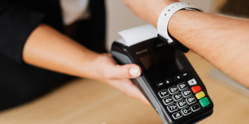 Tuvplānā redzams, kā cilvēks ar baltu viedpulksteni uz rokas veic bezkontakta maksājumu, pieliekot to pie melna POS termināļa, ko tur cits cilvēks.