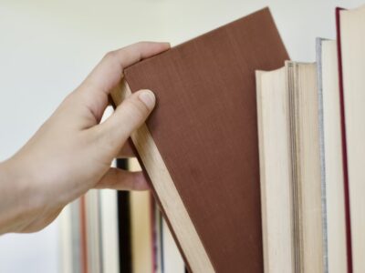 Cilvēka roka izvelk brūnu grāmatu no grāmatu plaukta, kurā redzamas arī citas grāmatas.