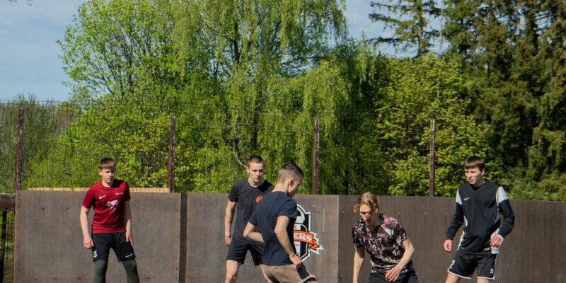 Pieci jauni vīrieši spēlē ielu futbolu uz āra asfalta laukuma ar koka sienām. Ēdolē notiks kausa izcīņa ielu futbolā